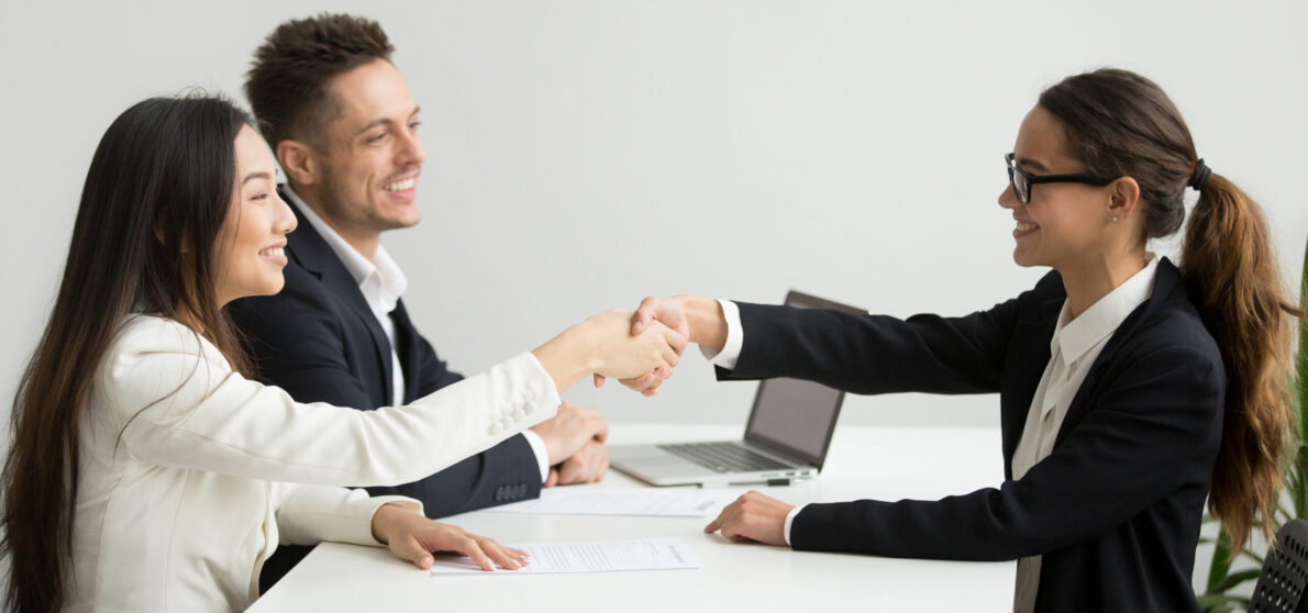 Smiling diverse businesswomen shake hands at group meeting, friendly asian hr handshaking congratulating hired applicant at job interview, satisfied millennial partners make contract deal concept