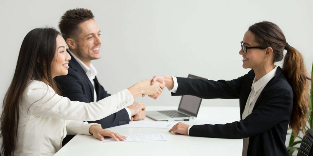 Smiling diverse businesswomen shake hands at group meeting, friendly asian hr handshaking congratulating hired applicant at job interview, satisfied millennial partners make contract deal concept